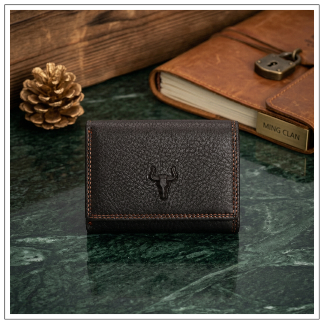 Front view of a MING CLAN dark brown top-grain leather trifold wallet with an embossed bull logo, resting on a green marble surface next to a vintage leather journal.