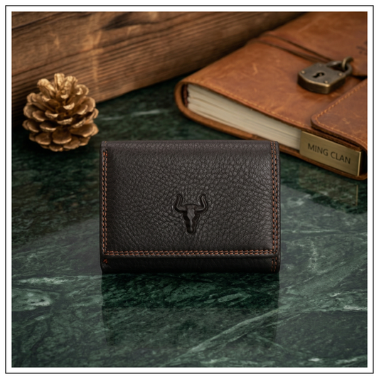 Front view of a MING CLAN dark brown top-grain leather trifold wallet with an embossed bull logo, resting on a green marble surface next to a vintage leather journal.